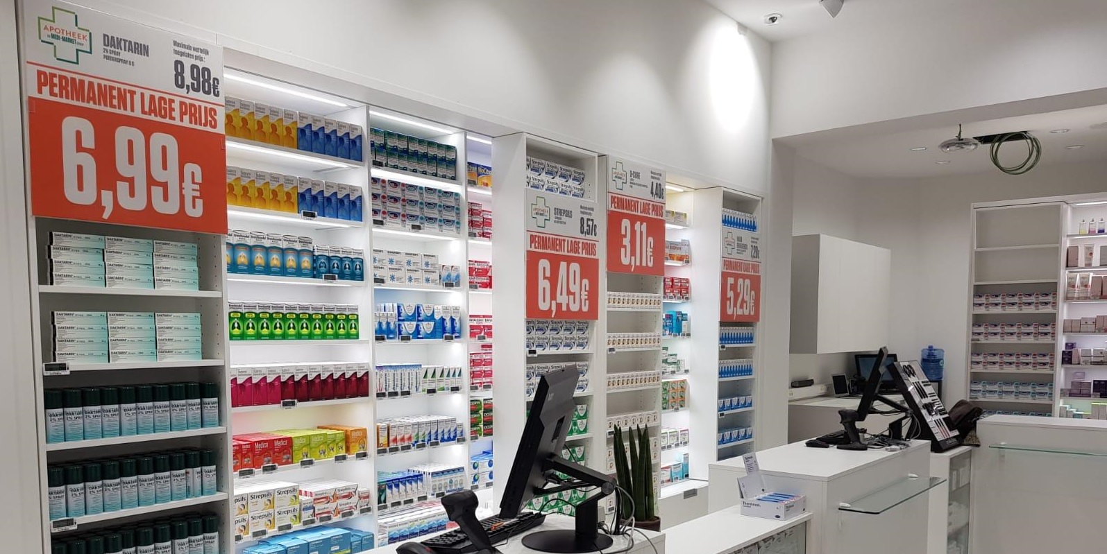 Rayons d'une pharmacie avec des produits en boîtes colorées et étiquettes de prix en rouge, avec un comptoir et un écran d'ordinateur au premier plan.