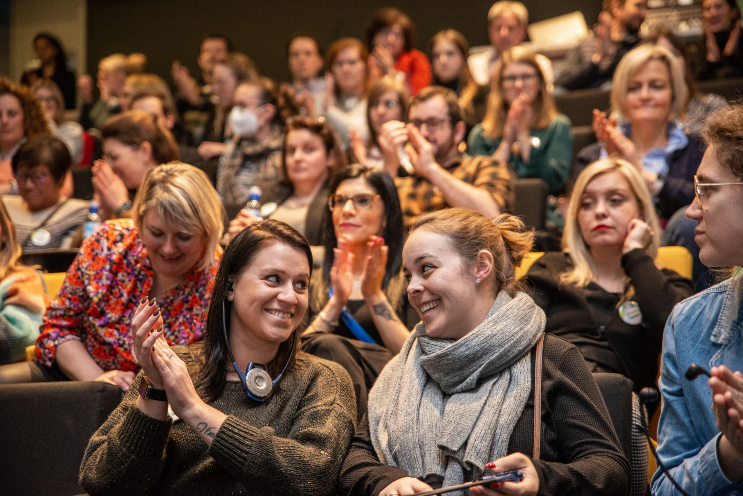 Un groupe de personnes assises dans un auditorium, certaines applaudissant, deux femmes souriantes au premier plan.
