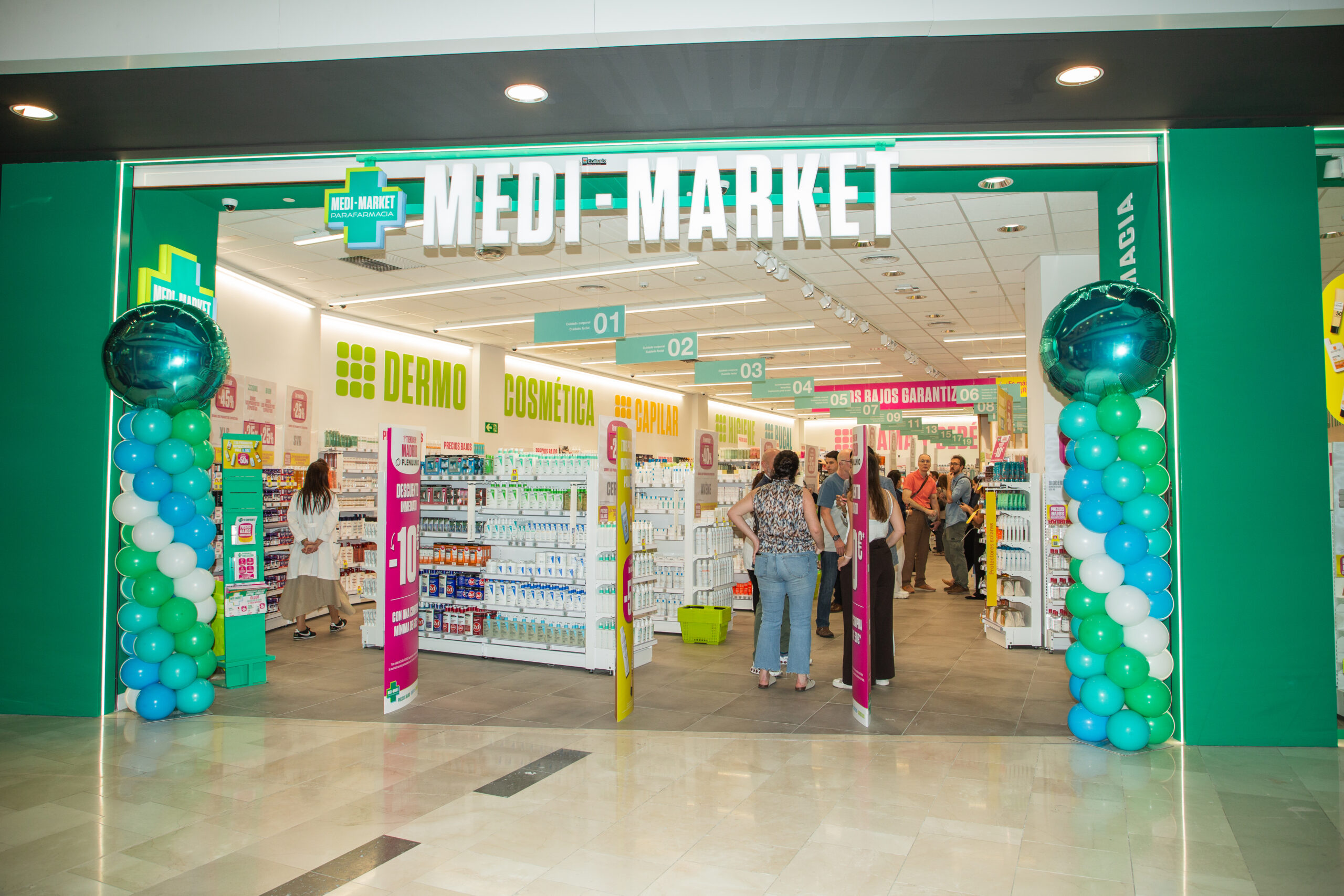 Entrée d'un magasin Medi-Market avec des ballons verts et blancs, des rayons de produits cosmétiques et des clients à l'intérieur.