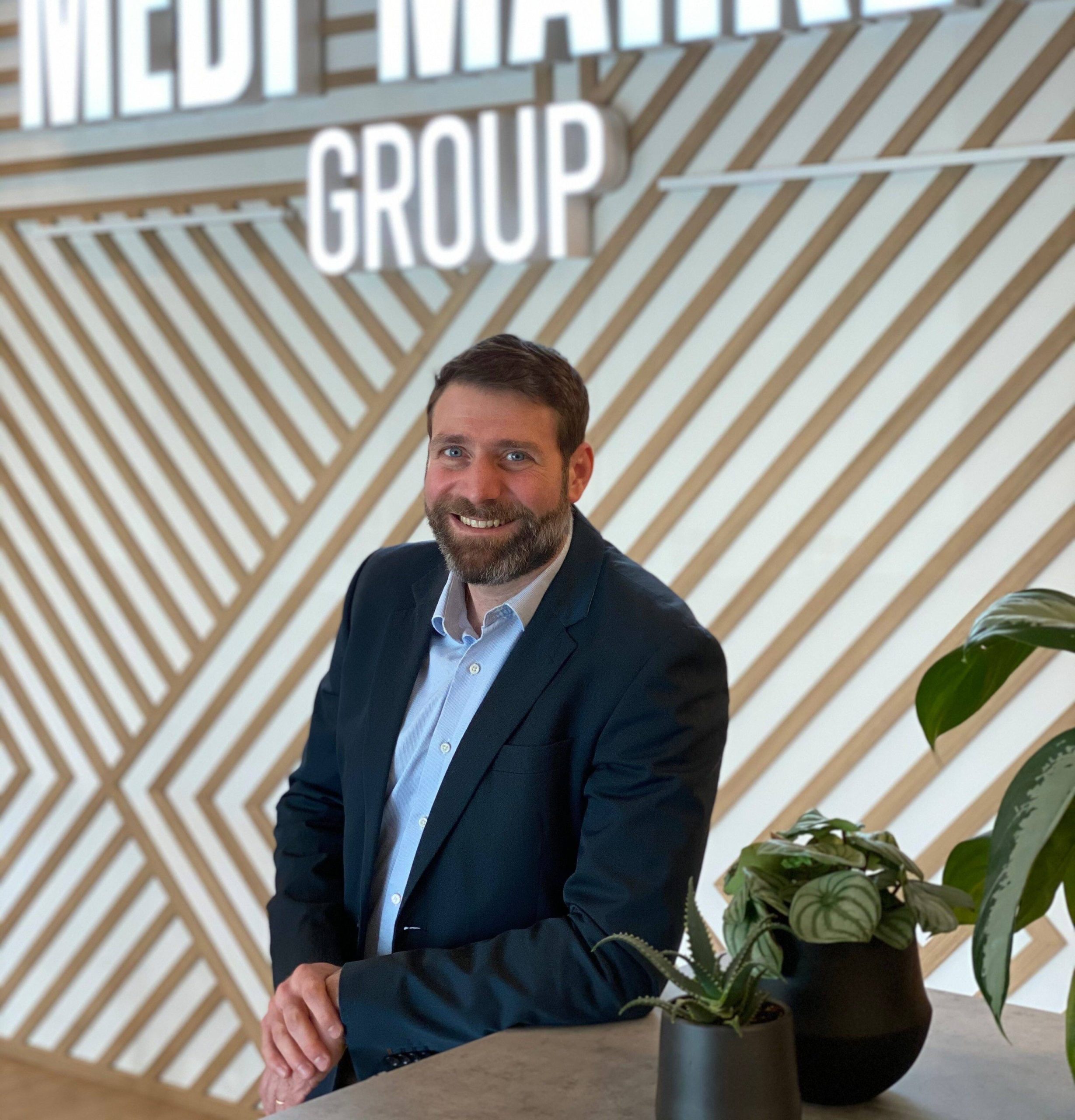 Un homme en costume bleu pose devant un panneau "MEDI MARKET GROUP" avec des plantes vertes en pot au premier plan.