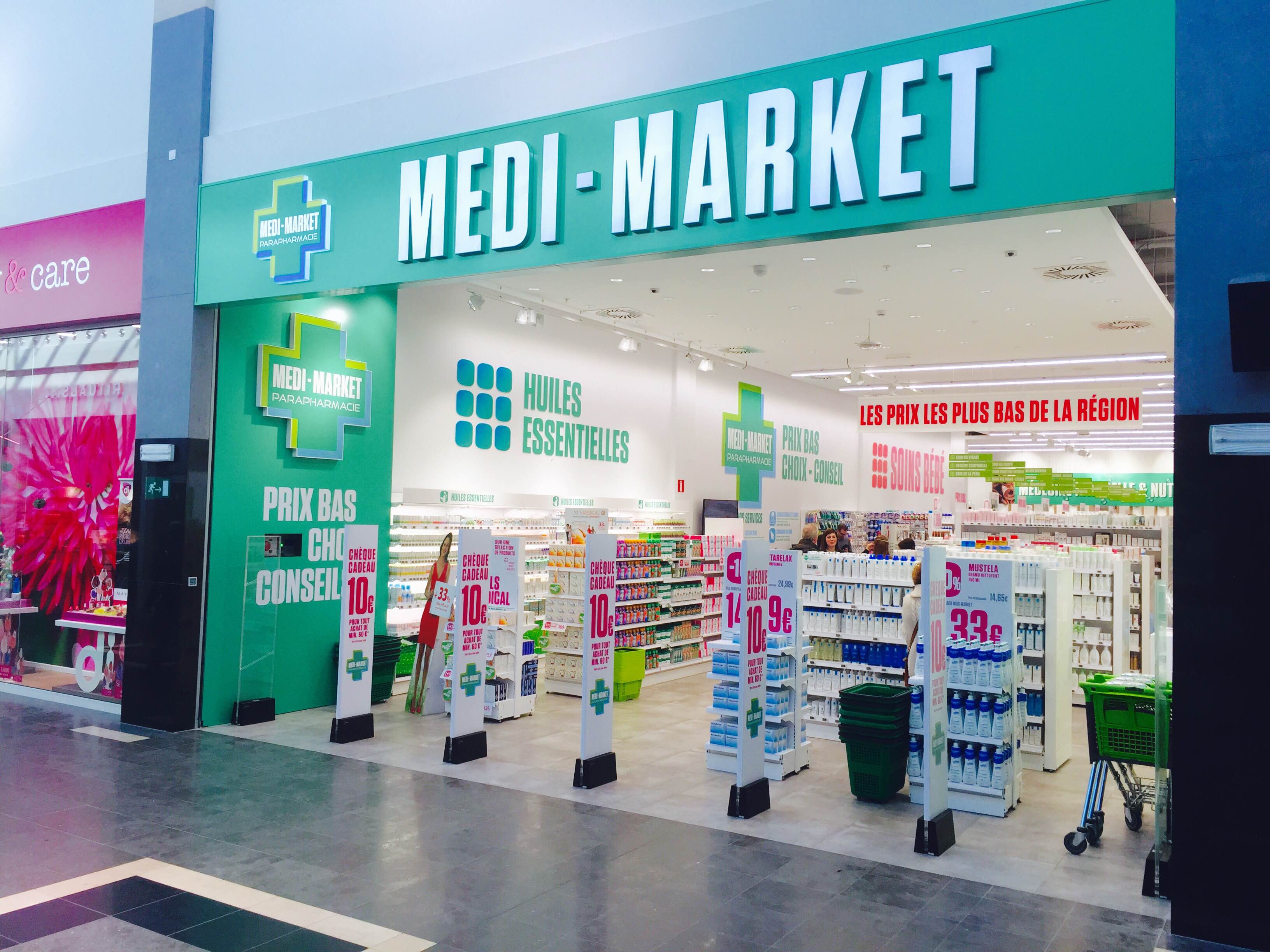 Façade d'une parapharmacie Medi-Market avec enseigne verte, étagères de produits, affiches promotionnelles et chariots verts à l'entrée.