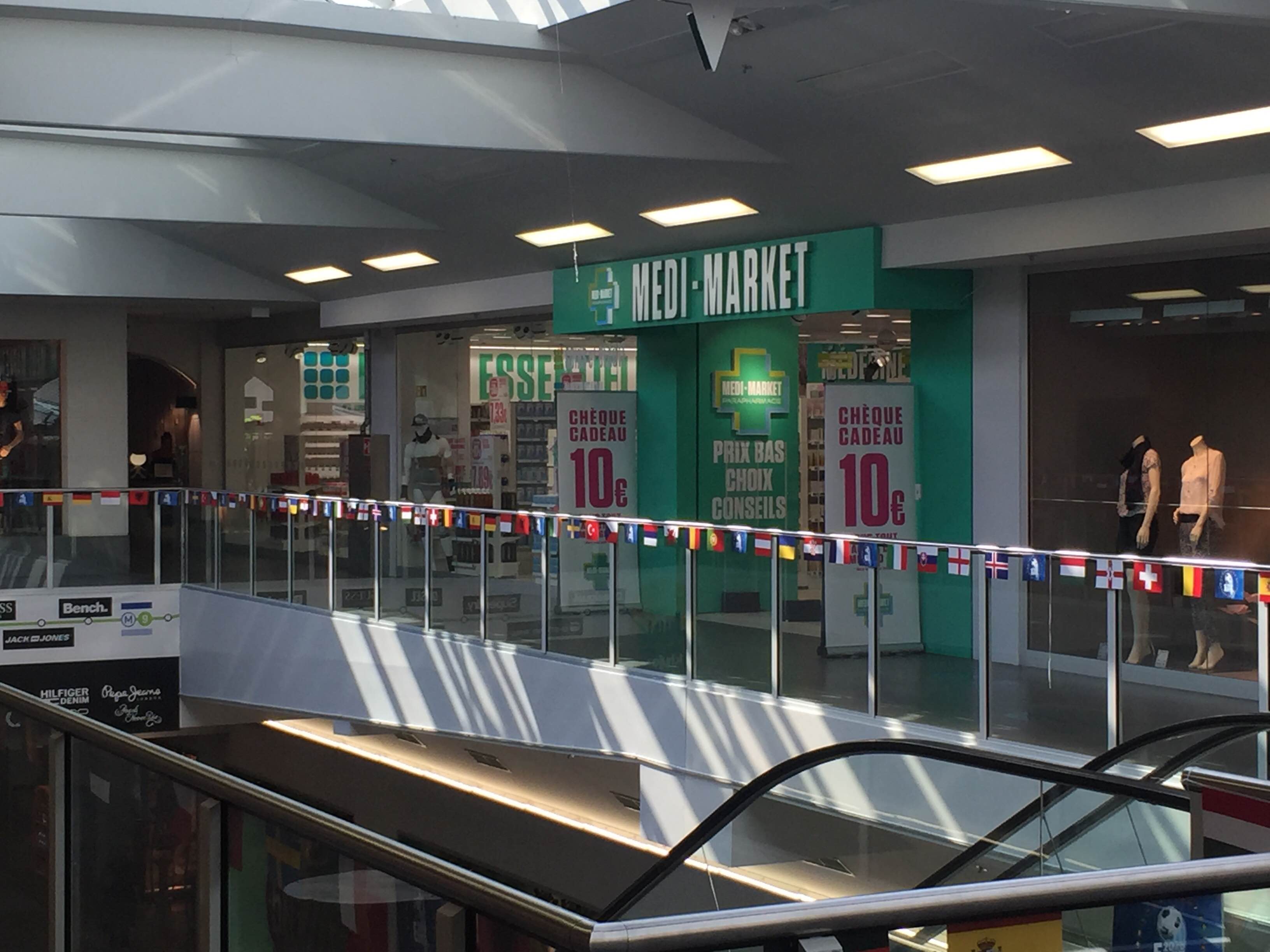 Façade de magasin Medi-Market avec mannequins en vitrine et guirlande de drapeaux internationaux suspendue au-dessus d'un escalator.