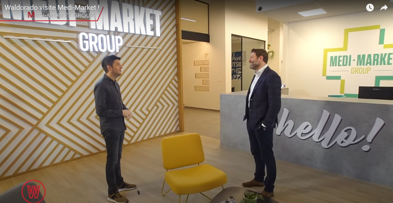 Deux hommes discutent dans un hall d'accueil moderne avec un mur décoratif, un fauteuil jaune et un comptoir portant l'inscription "hello!".