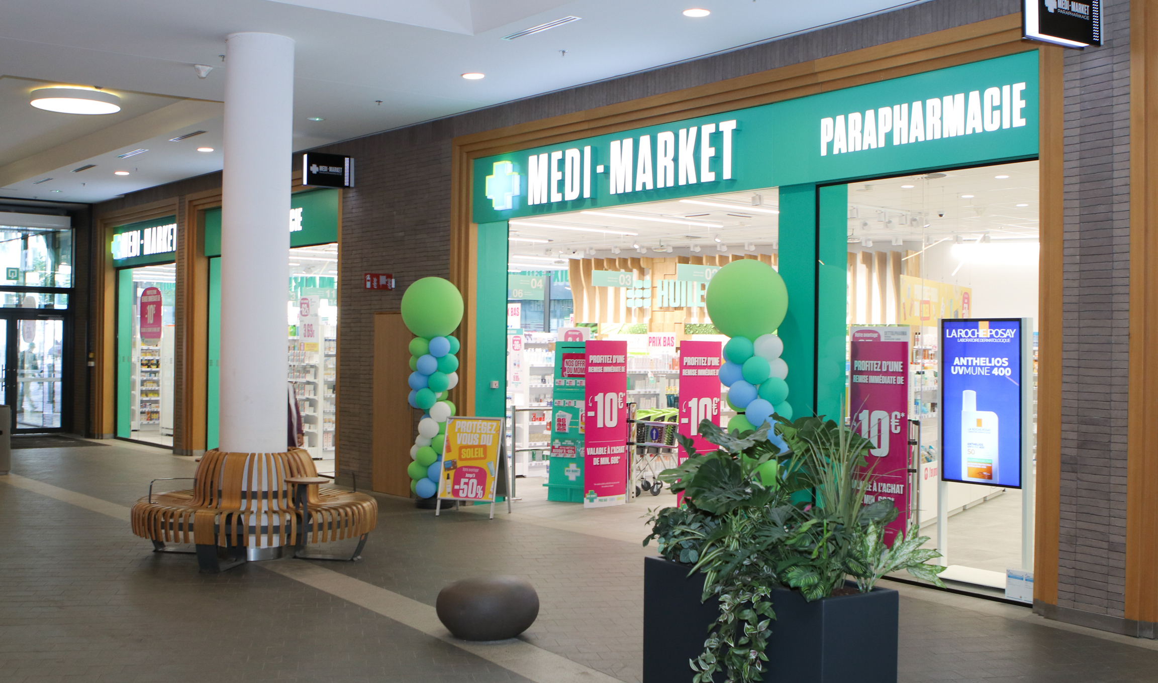 Entrée de la parapharmacie Medi-Market avec ballons verts et bleus, affiches promotionnelles et présentoirs de produits visibles à l'intérieur.