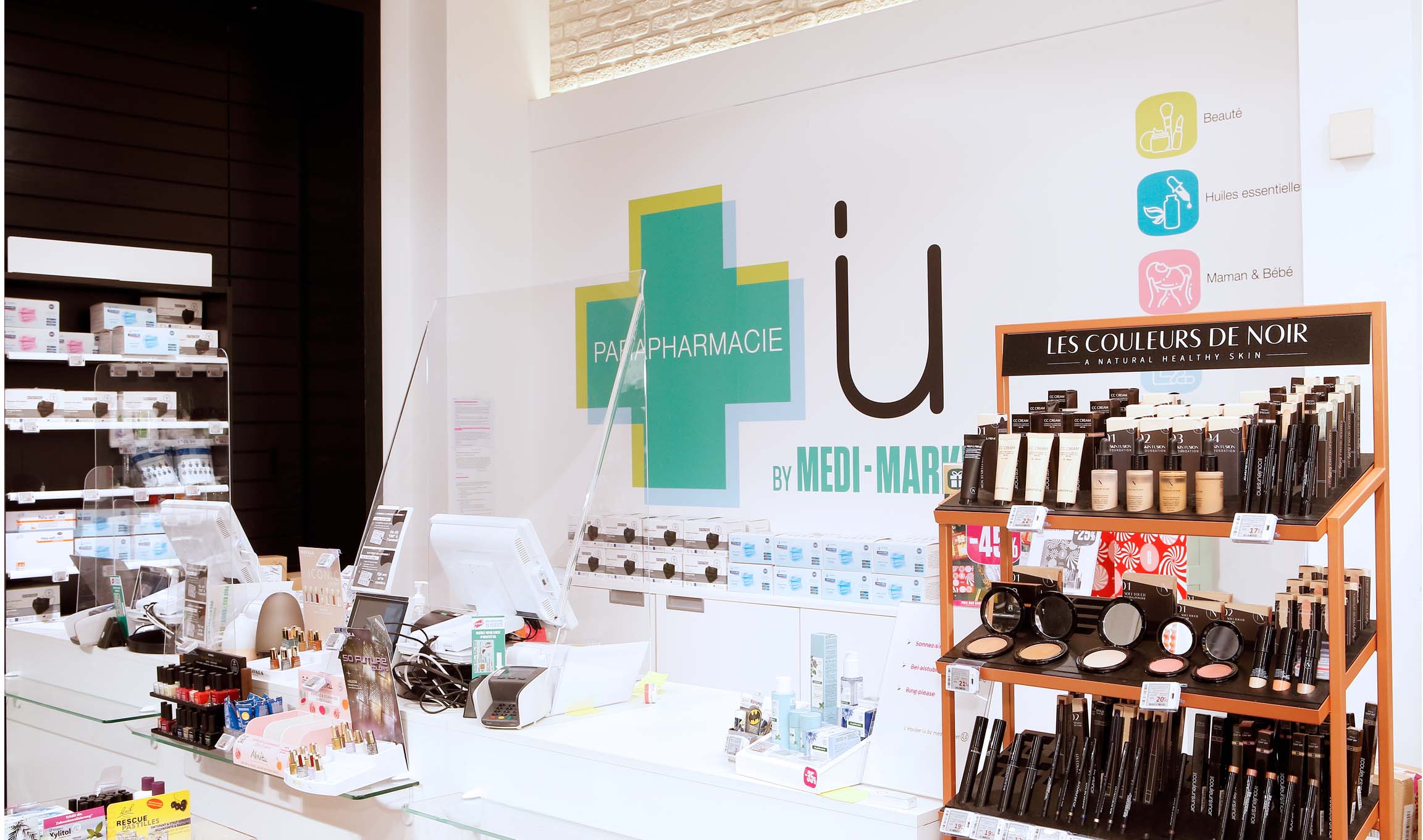 Comptoir de parapharmacie avec produits de beauté, affiches et caisse enregistreuse, présentoir de cosmétiques "Les Couleurs de Noir" à droite.