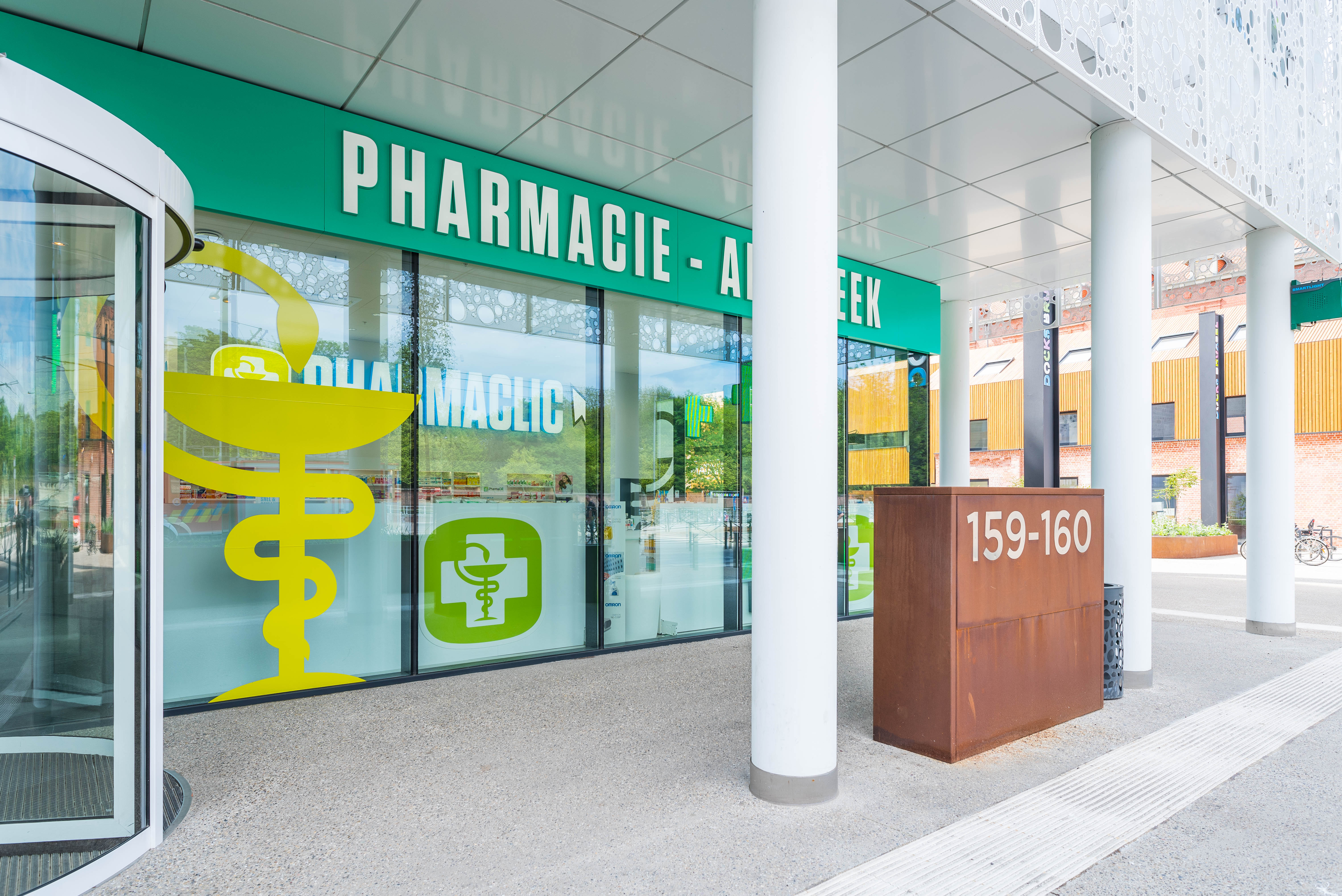Façade d'une pharmacie avec enseigne verte, symbole médical jaune et numéro 159-160 sur une boîte en métal rouillé.
