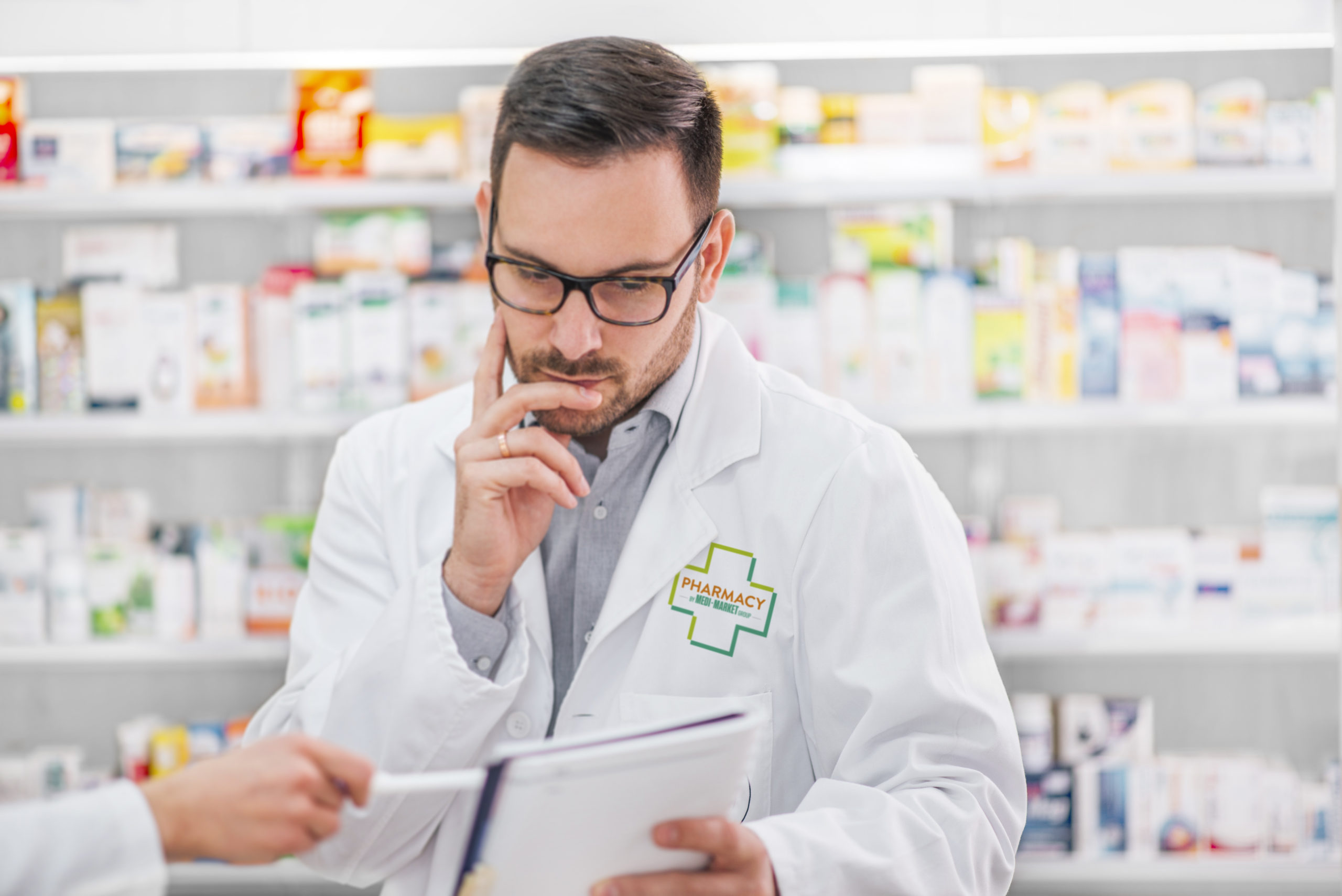 Un homme en blouse blanche lit un document dans une pharmacie, avec des étagères remplies de produits en arrière-plan.