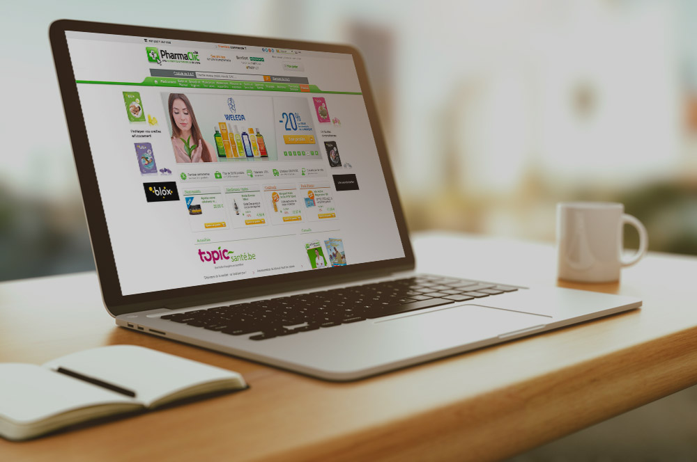Ordinateur portable affichant une page web de pharmacie en ligne sur un bureau en bois, avec un carnet et une tasse blanche à côté.
