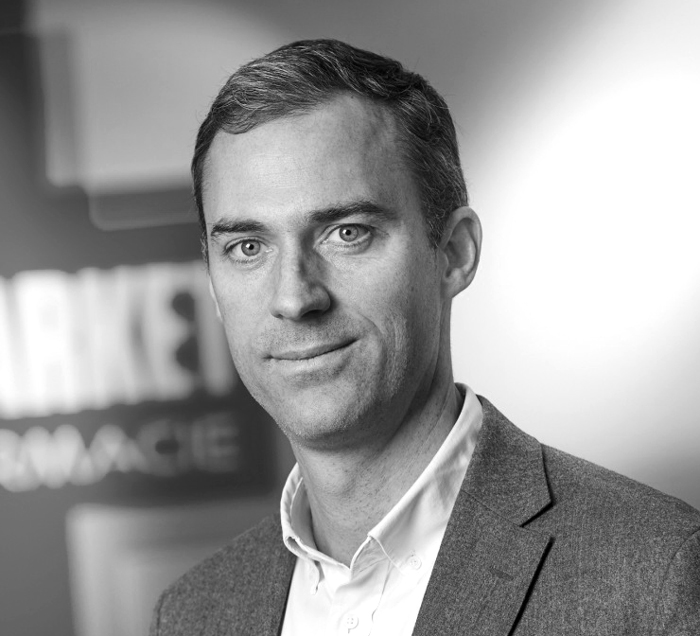 Un homme en costume gris et chemise blanche pose devant un fond flou avec des lettres visibles.