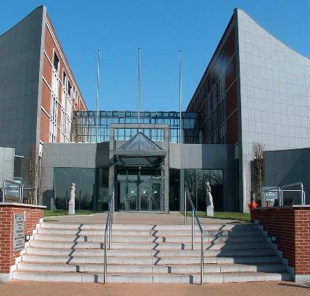 Bâtiment moderne avec façade en verre et briques rouges, escalier central menant à l'entrée principale entourée de statues.