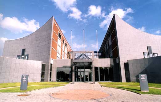 Bâtiment moderne en béton gris avec façades incurvées, trois mâts sans drapeaux devant l'entrée vitrée, sous un ciel partiellement nuageux.