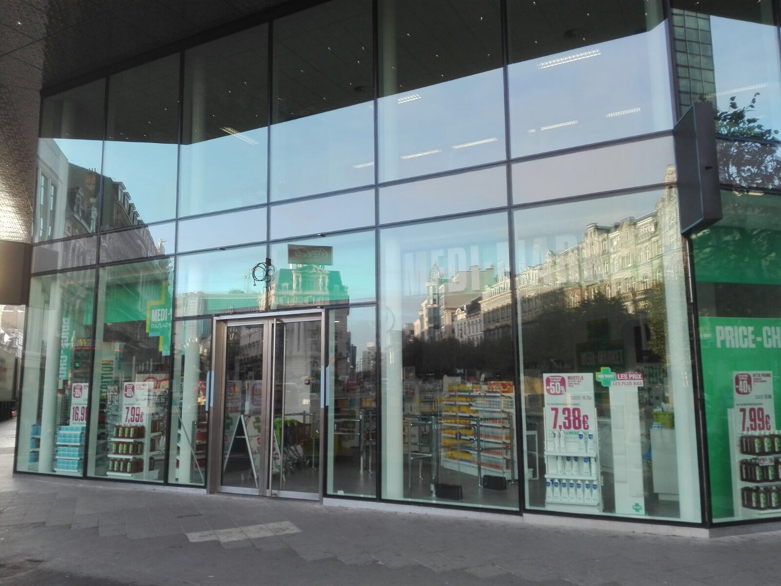 Façade en verre d'une pharmacie avec reflets urbains, affiches de prix et étagères de produits visibles à l'intérieur.