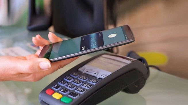 Une main utilisant un smartphone noir pour effectuer un paiement sans contact sur un terminal de carte bancaire gris.