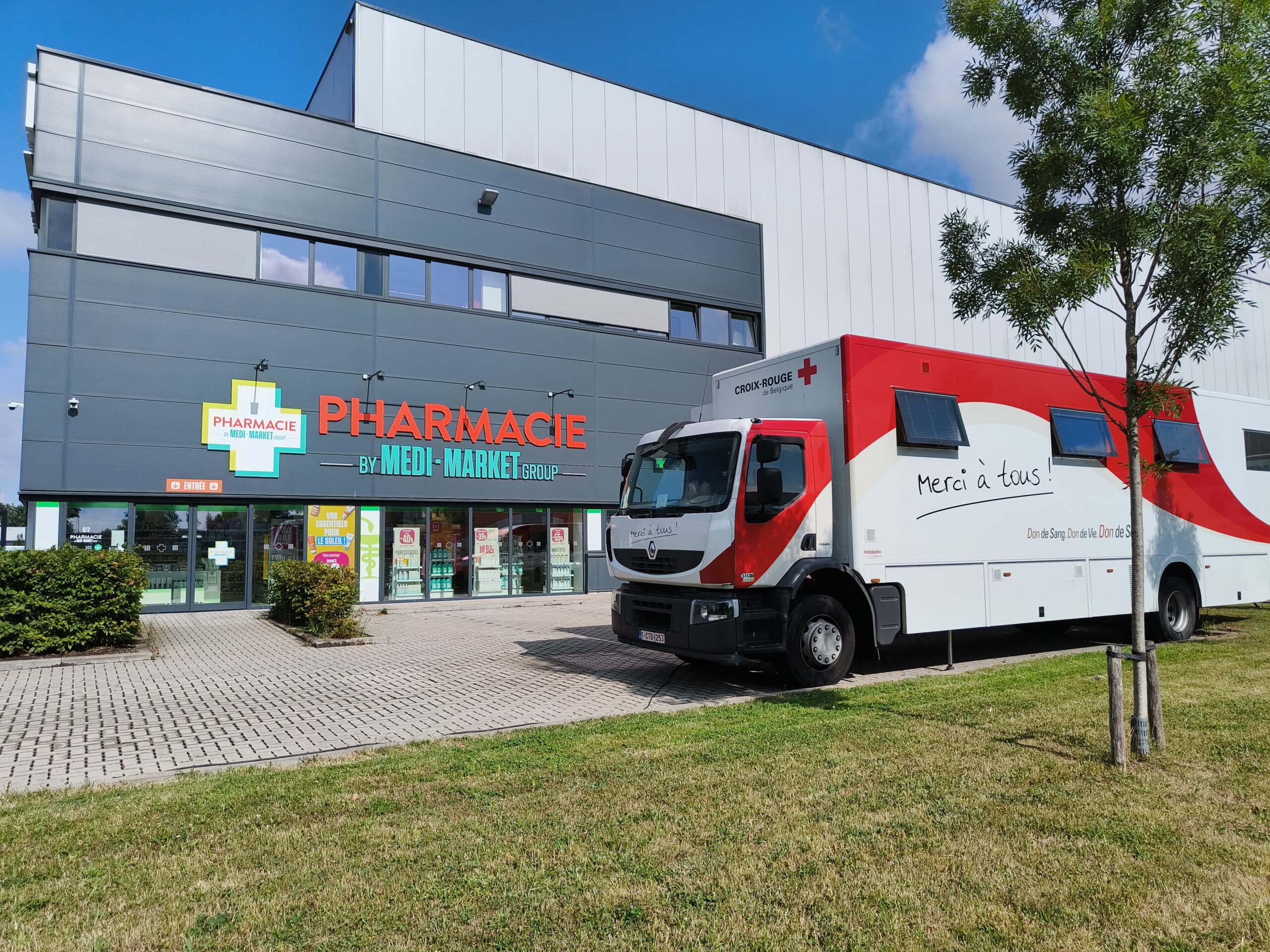 Un camion blanc et rouge de la Croix-Rouge est garé devant une pharmacie moderne, sur un trottoir pavé entouré de verdure.