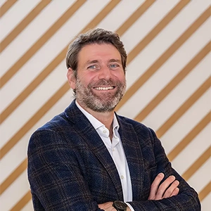 Un homme en costume à carreaux bleus sourit devant un mur à rayures diagonales beiges et blanches.