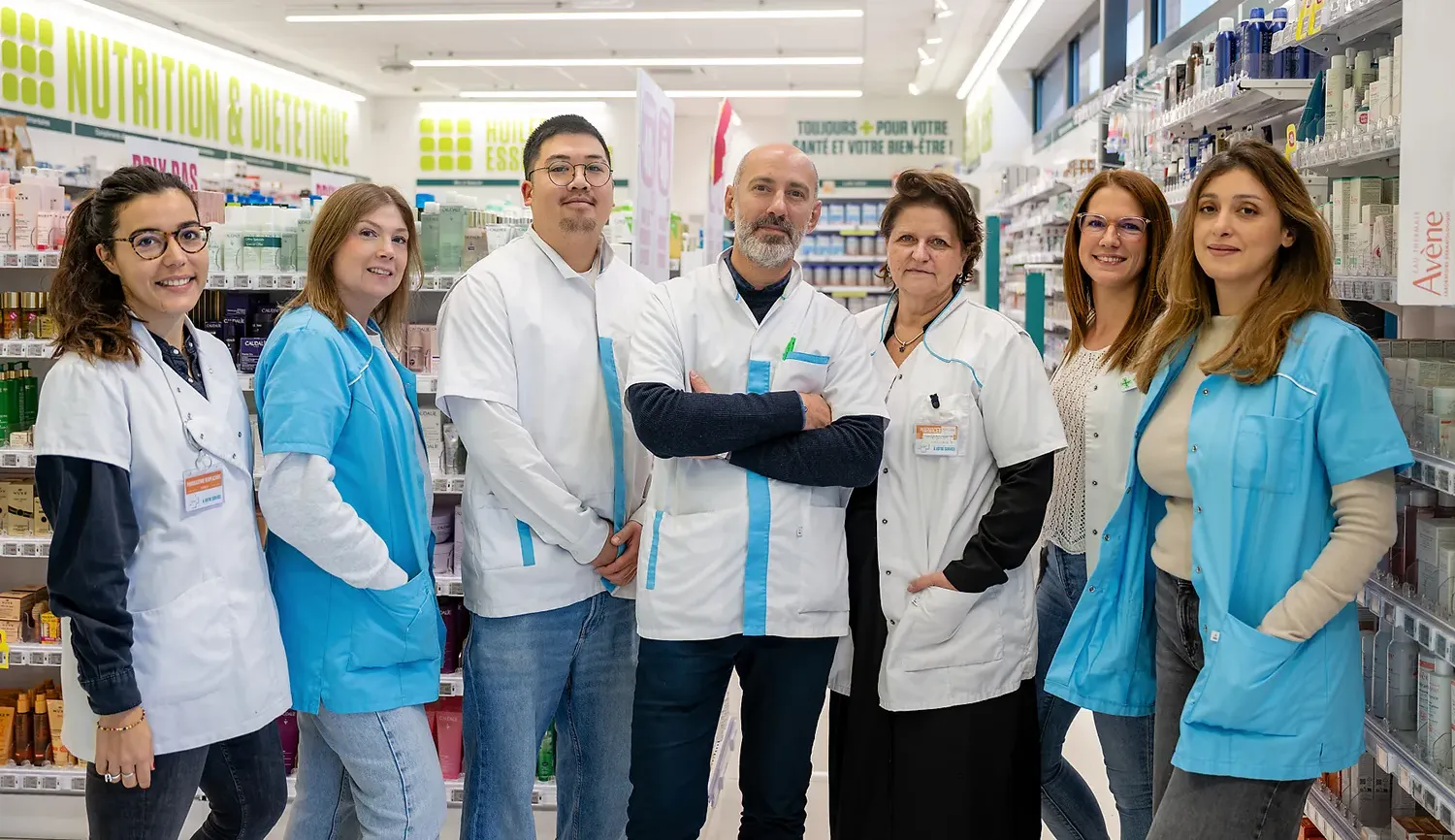 Un groupe de sept personnes en blouses blanches et bleues se tient dans une pharmacie, entouré d'étagères de produits.